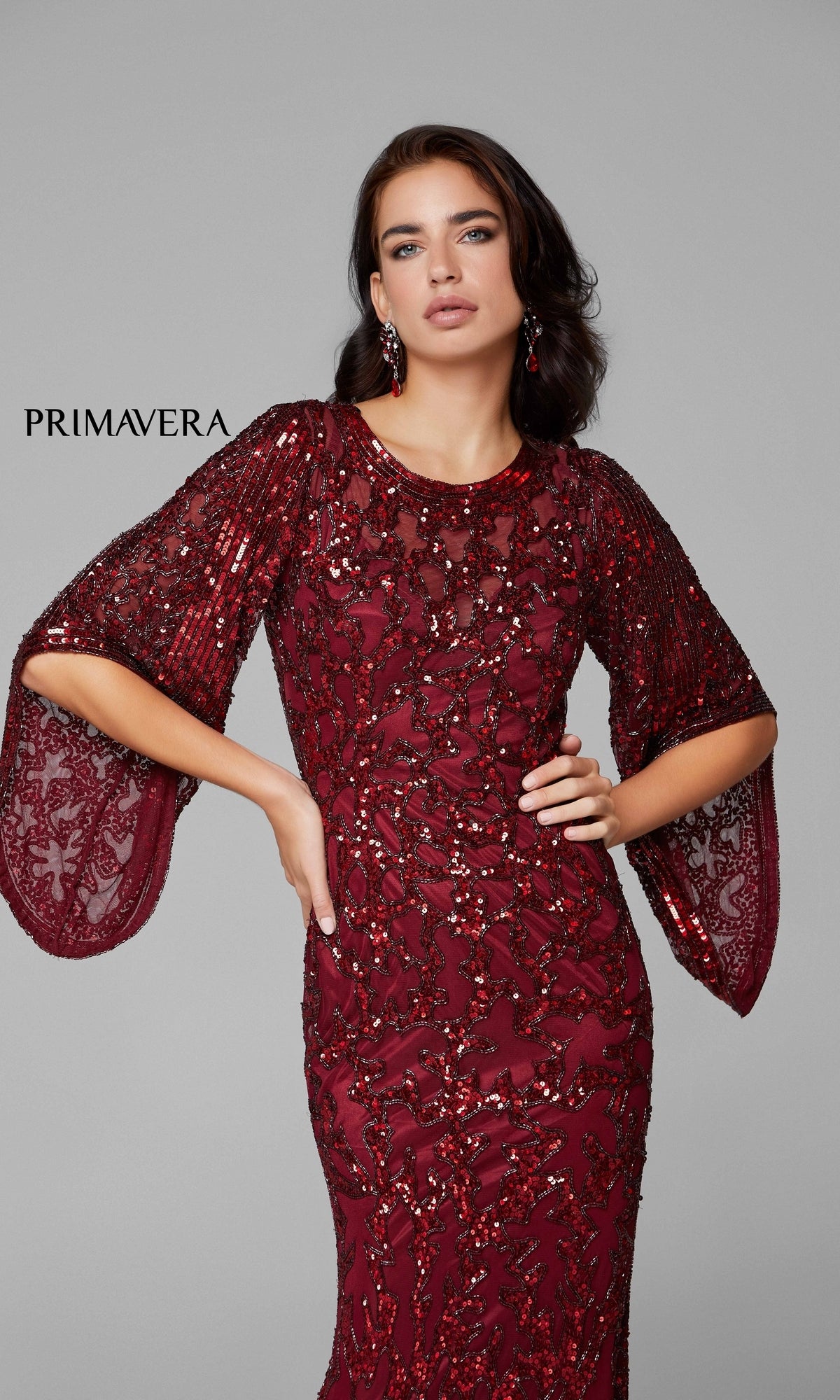 A woman models the Long Bell Sleeve Beaded Evening Gown 9713 in burgundy, featuring elegant lace bell sleeves, against a plain gray background.